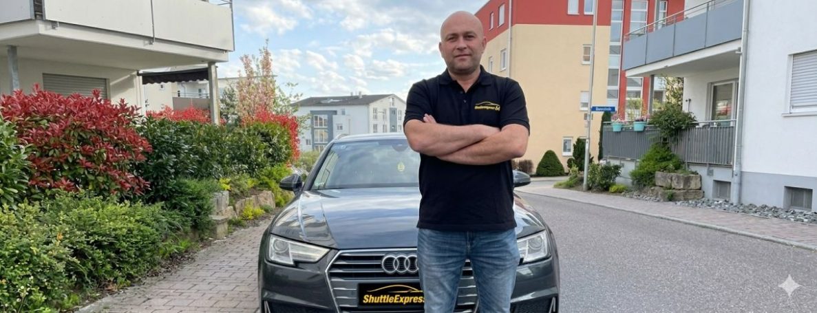 Mann steht mit verschränkten Armen vor grauem Auto auf einer Straße.