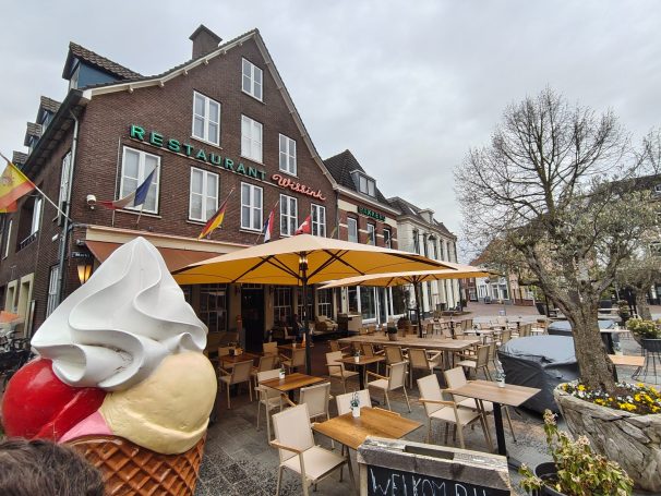 Eisbecher mit drei Kugeln vor einem Café mit Sonnenschirmen und Tischen in einer Stadt.